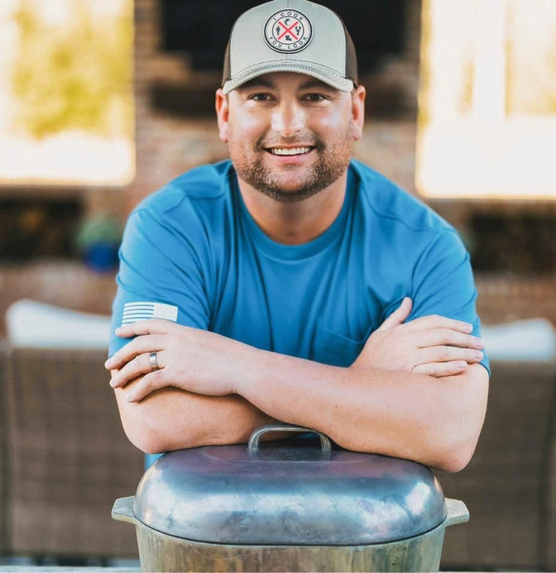 Austin Dennis with I Cook You Look, wearing company merch and a blue shirt, standing next to a barbecue grill outdoors. He was always born for the grill. 